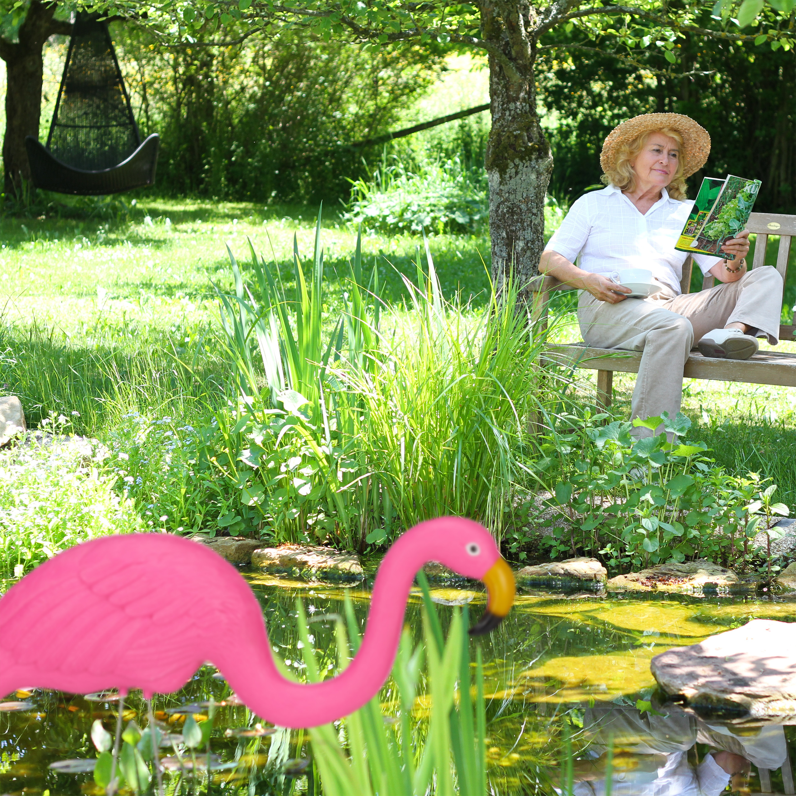 Eine Frau mit Strohhut liest auf einer Bank am sonnigen Gartenteich mit pinkem Flamingo eine Zeitschrift.
