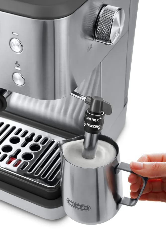 Una mano sostiene una jarra de leche de acero inoxidable bajo el vaporizador de una cafetera DeLonghi.