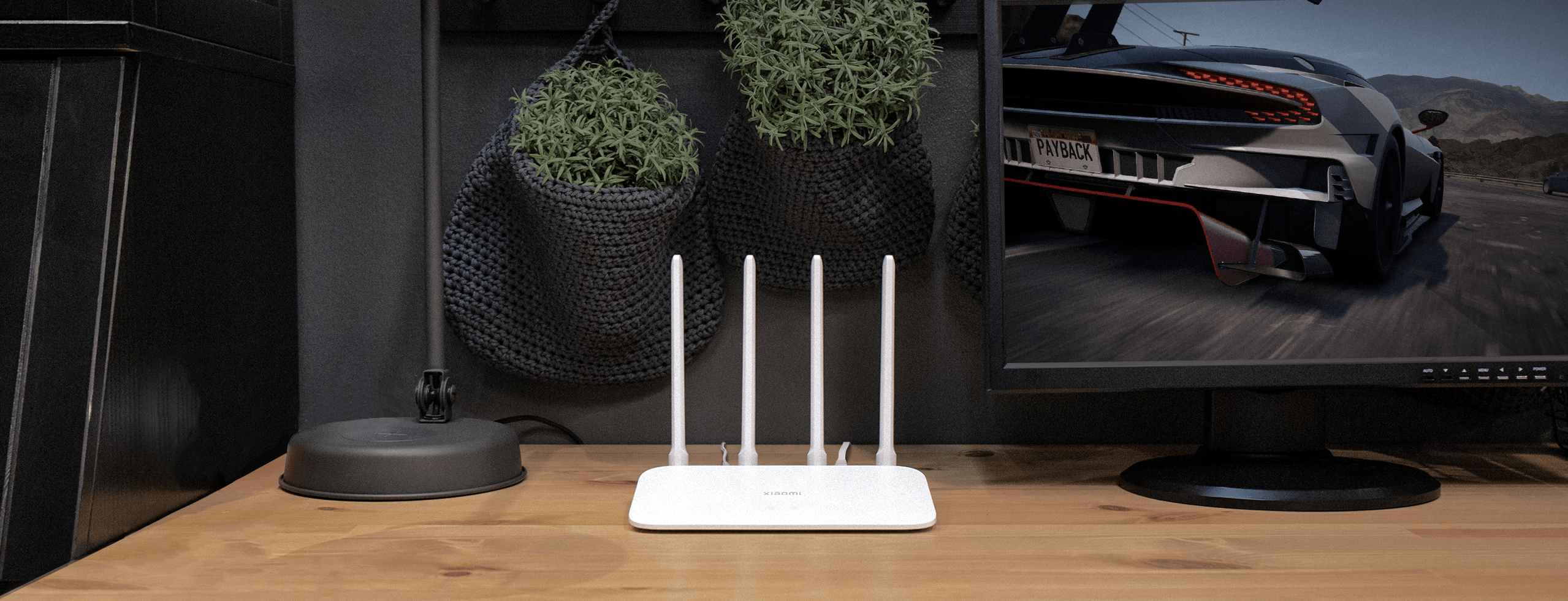 Un router Xiaomi blanco con cuatro antenas se encuentra en un escritorio de madera junto a un monitor y plantas colgantes.