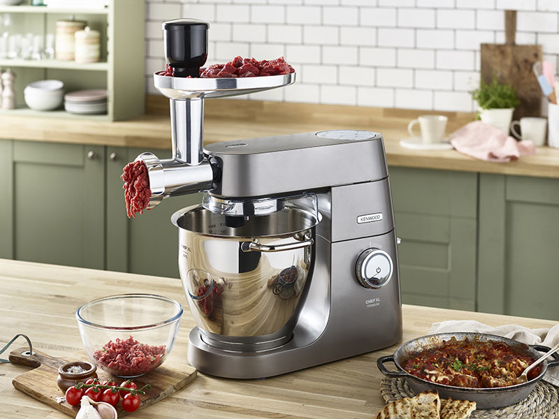 Un robot de cocina Kenwood plateado con accesorio picador de carne en una encimera de madera.