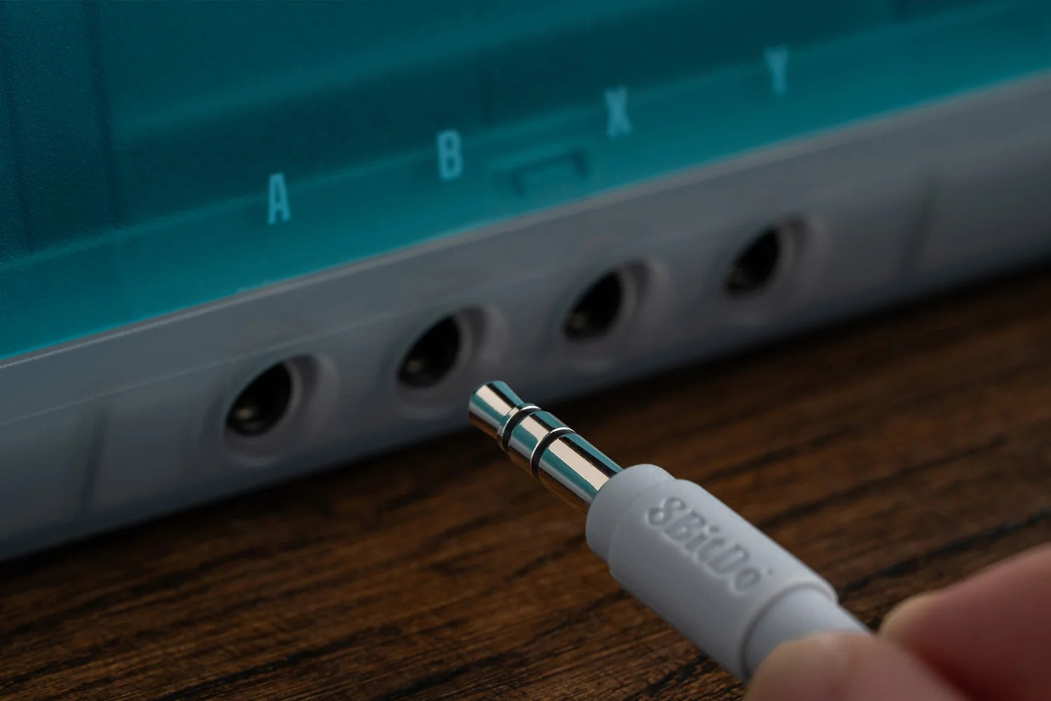 Ręka trzyma biały kabel audio 8BitDo w pobliżu morskiego urządzenia z portami oznaczonymi A, B, X, Y.
