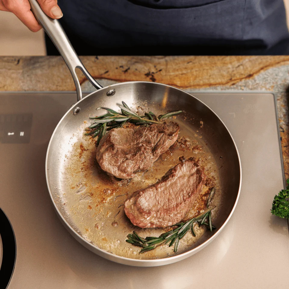 Two steaks and rosemary sprigs cook in a metal frying pan on a stovetop.