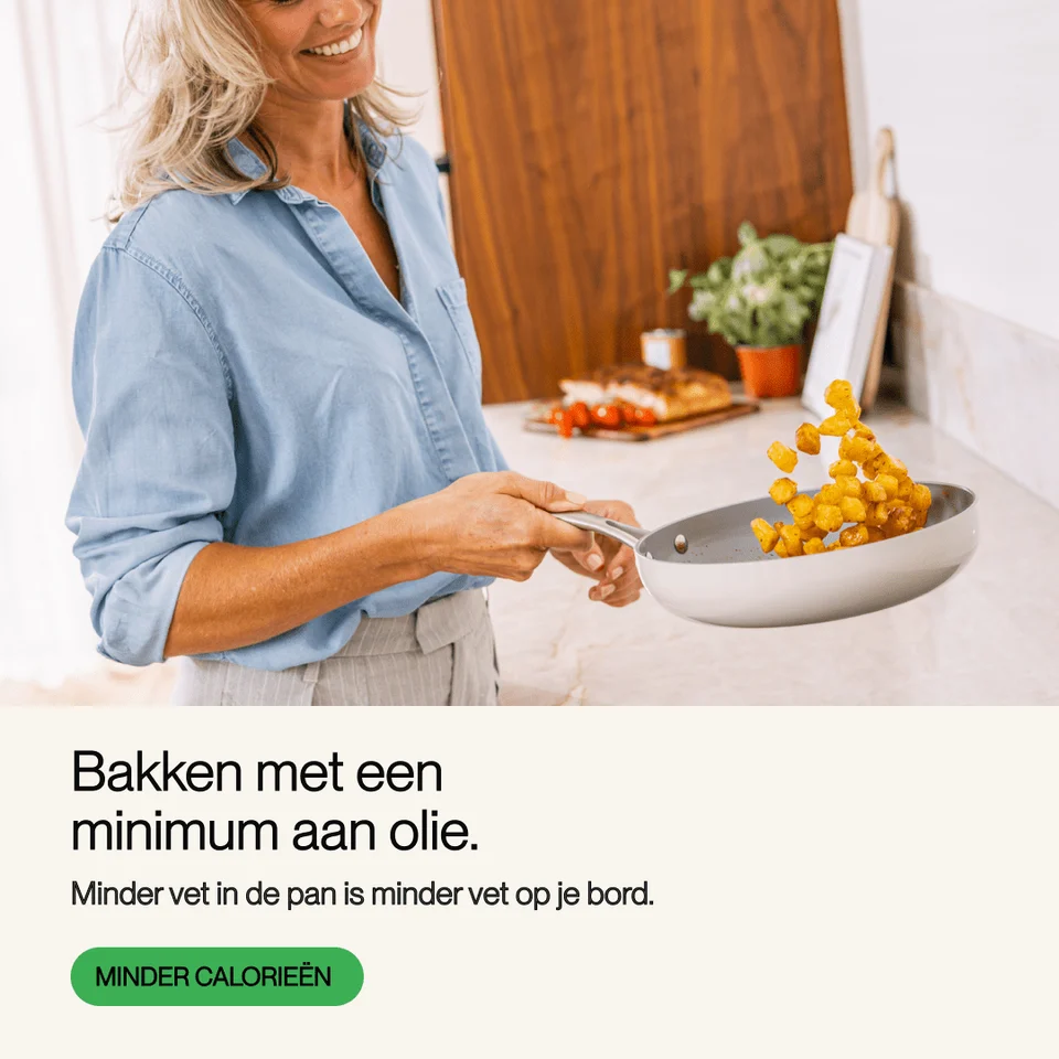 A smiling woman in a blue shirt flips golden potato cubes in a white frying pan in a bright kitchen.