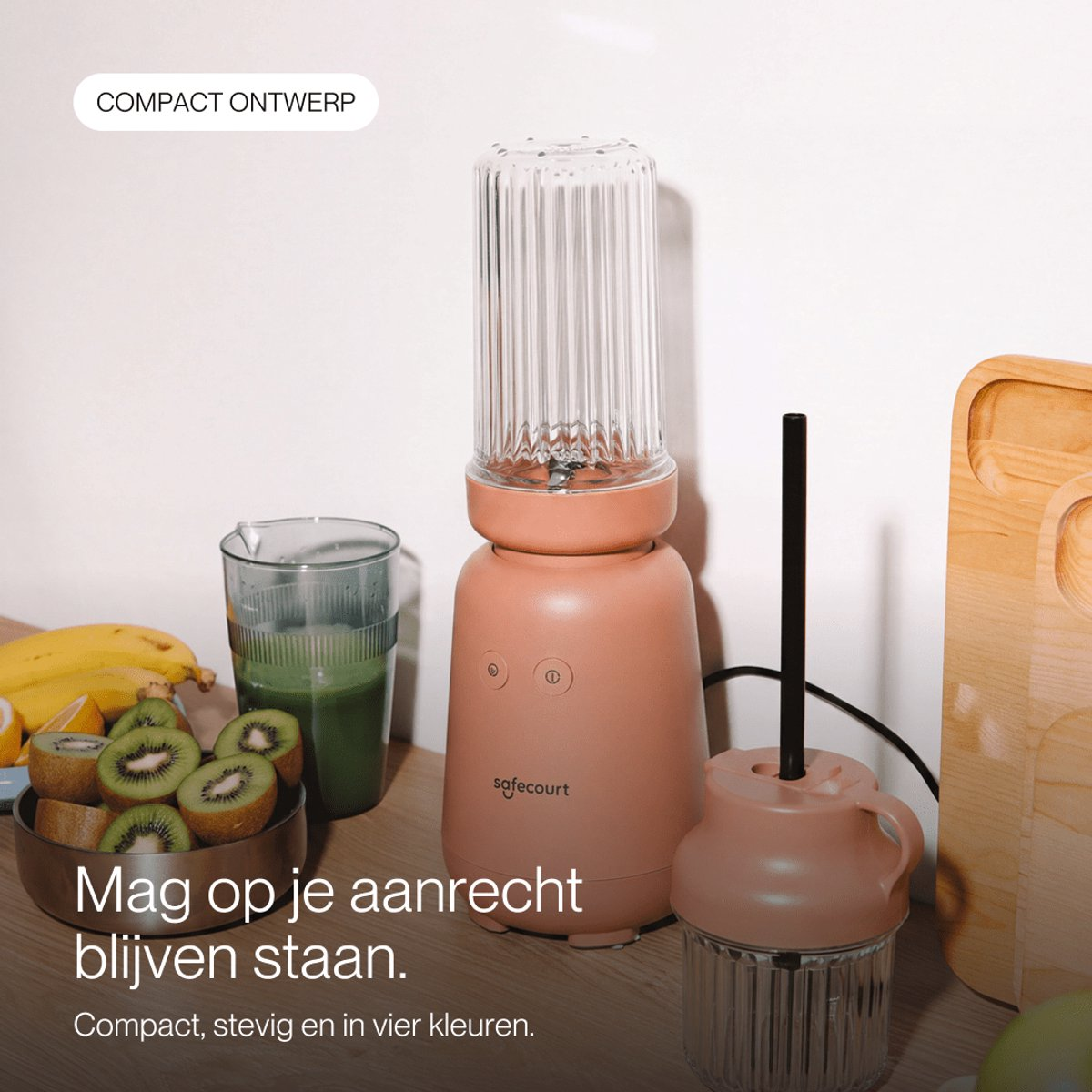 Un blender rose sur un comptoir en bois, avec des fruits coupés, un verre de smoothie vert et une planche à découper.