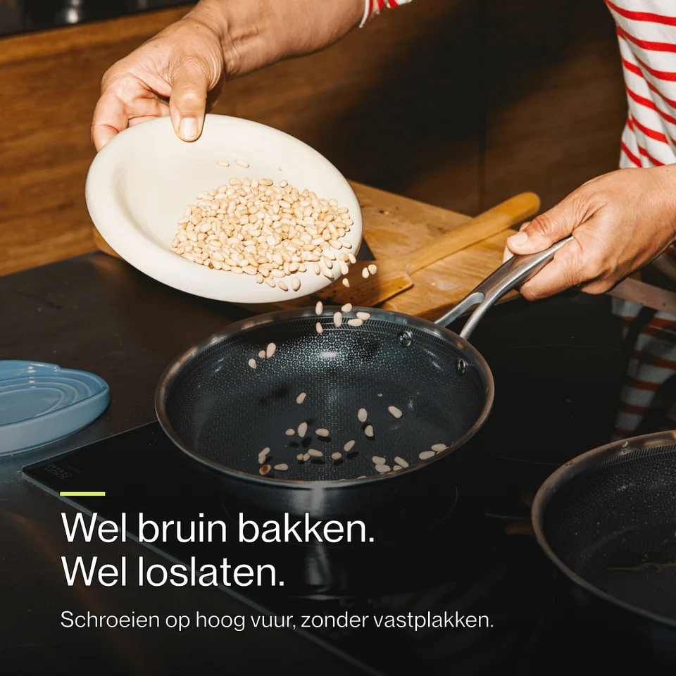 A person pours pine nuts from a white bowl into a black frying pan on a stovetop.