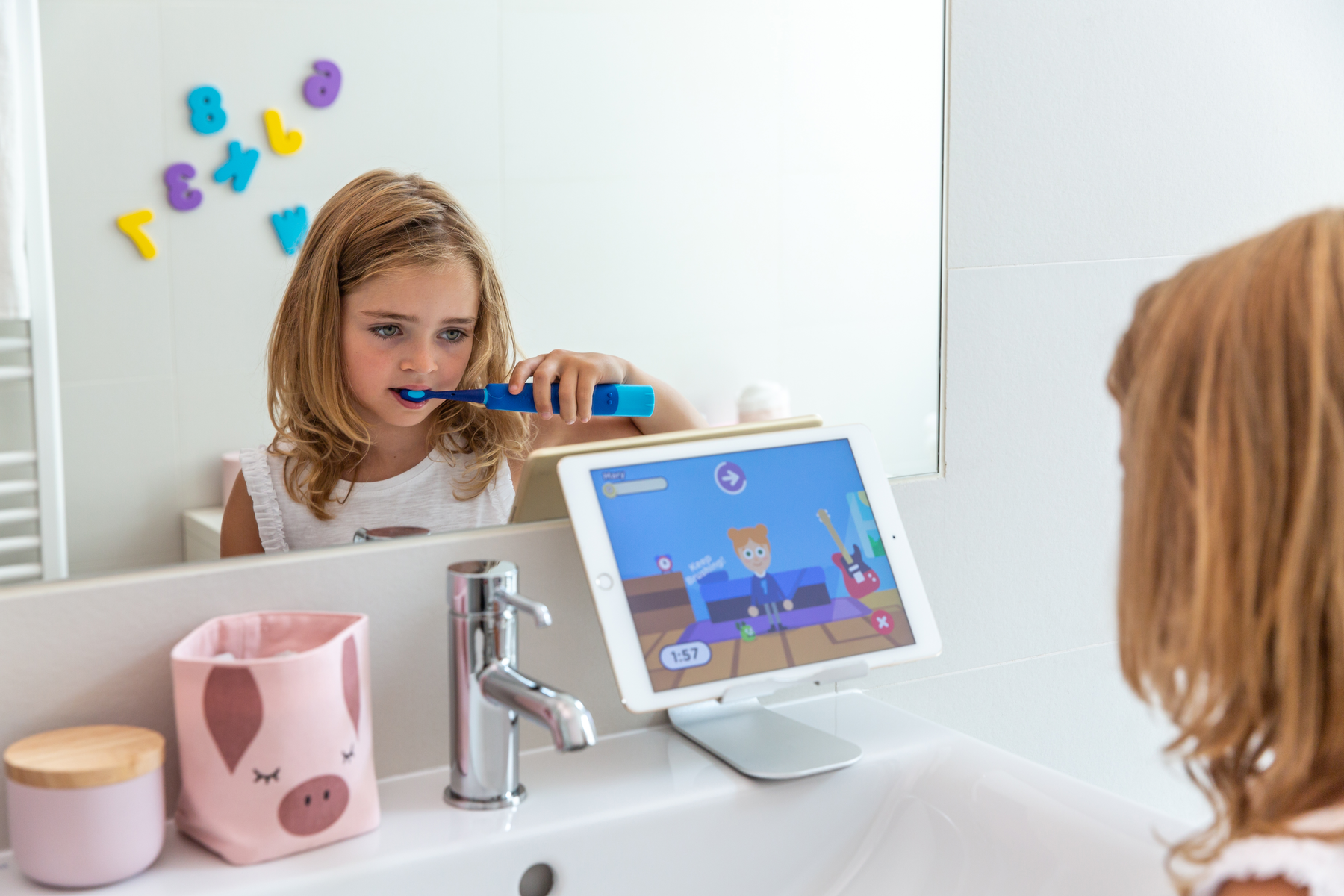 Una bambina bionda si lava i denti con uno spazzolino elettrico in bagno guardando un tablet.