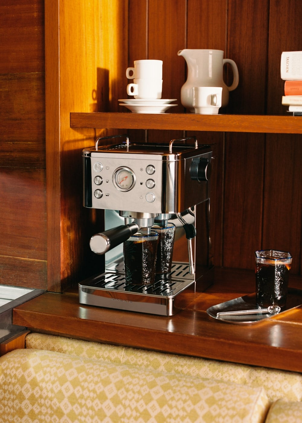 Una cafetera de acero inoxidable sobre una encimera de madera con vasos llenos y tazas blancas en el estante.