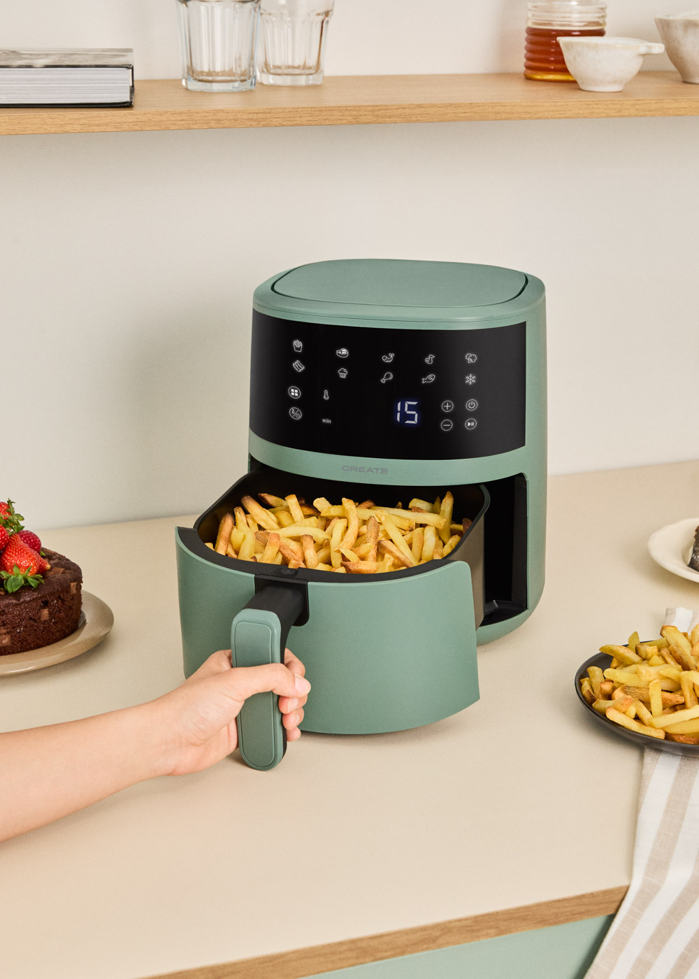 Una freidora de aire verde en un mostrador muestra una cesta llena de patatas fritas. Hay un pastel y patatas cerca.
