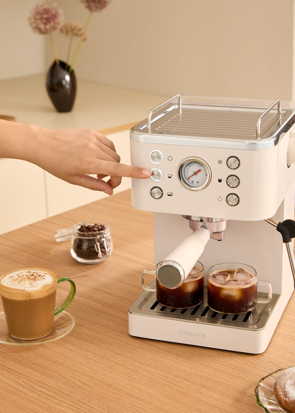 Una mano opera una cafetera blanca en una mesa de madera con capuchino y granos de café.