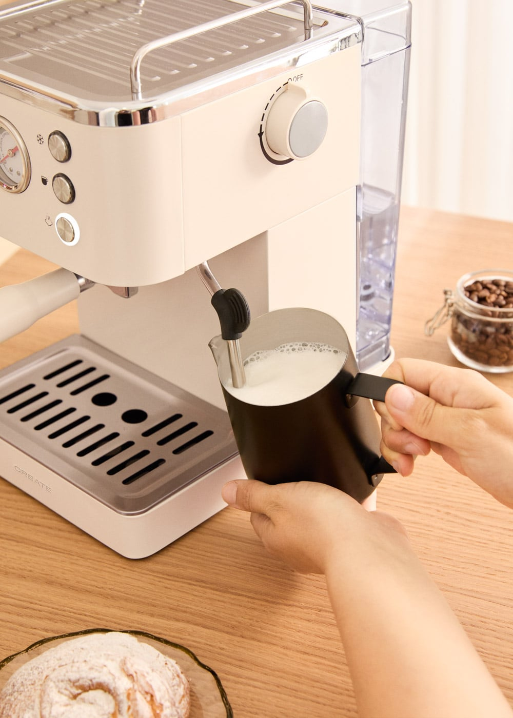 Una persona espuma leche en una jarra negra usando una máquina de espresso beige claro en una mesa de madera.