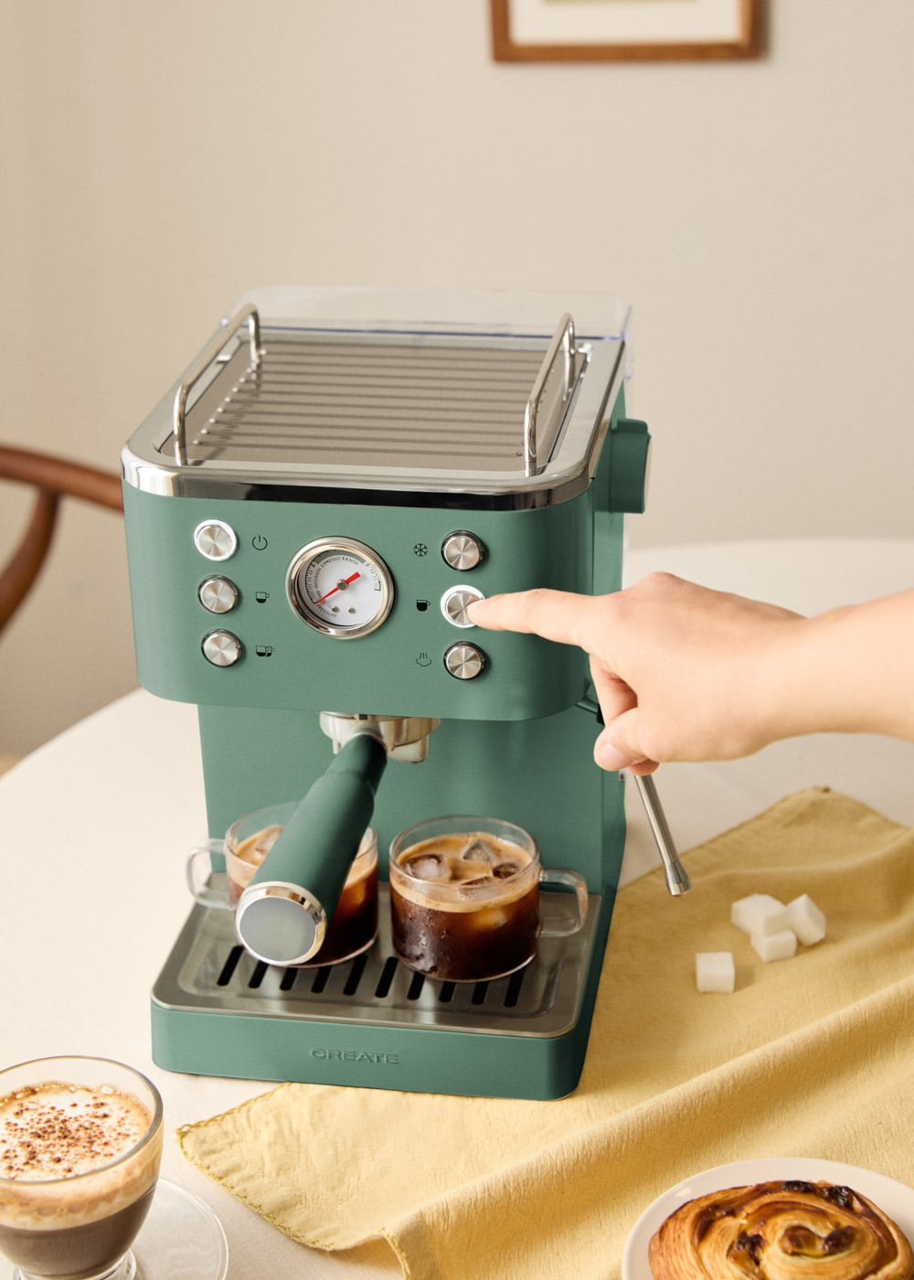 Una mano opera una cafetera espresso verde con dos tazas de café debajo y terrones de azúcar cerca.