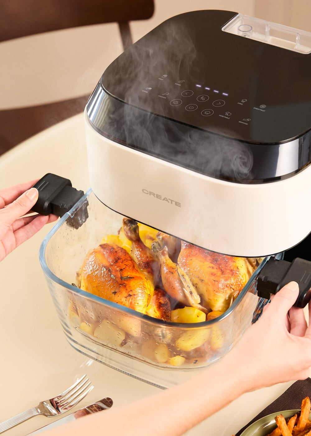 Manos sostienen la cesta de vidrio de una freidora de aire con pollo y patatas cocinados, con vapor saliendo.