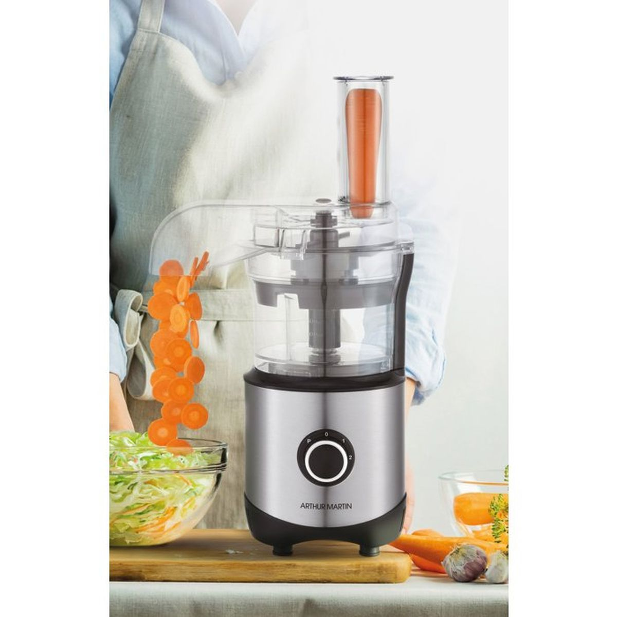 Un robot da cucina affetta le carote facendole cadere in una ciotola, con verdure sul tavolo.
