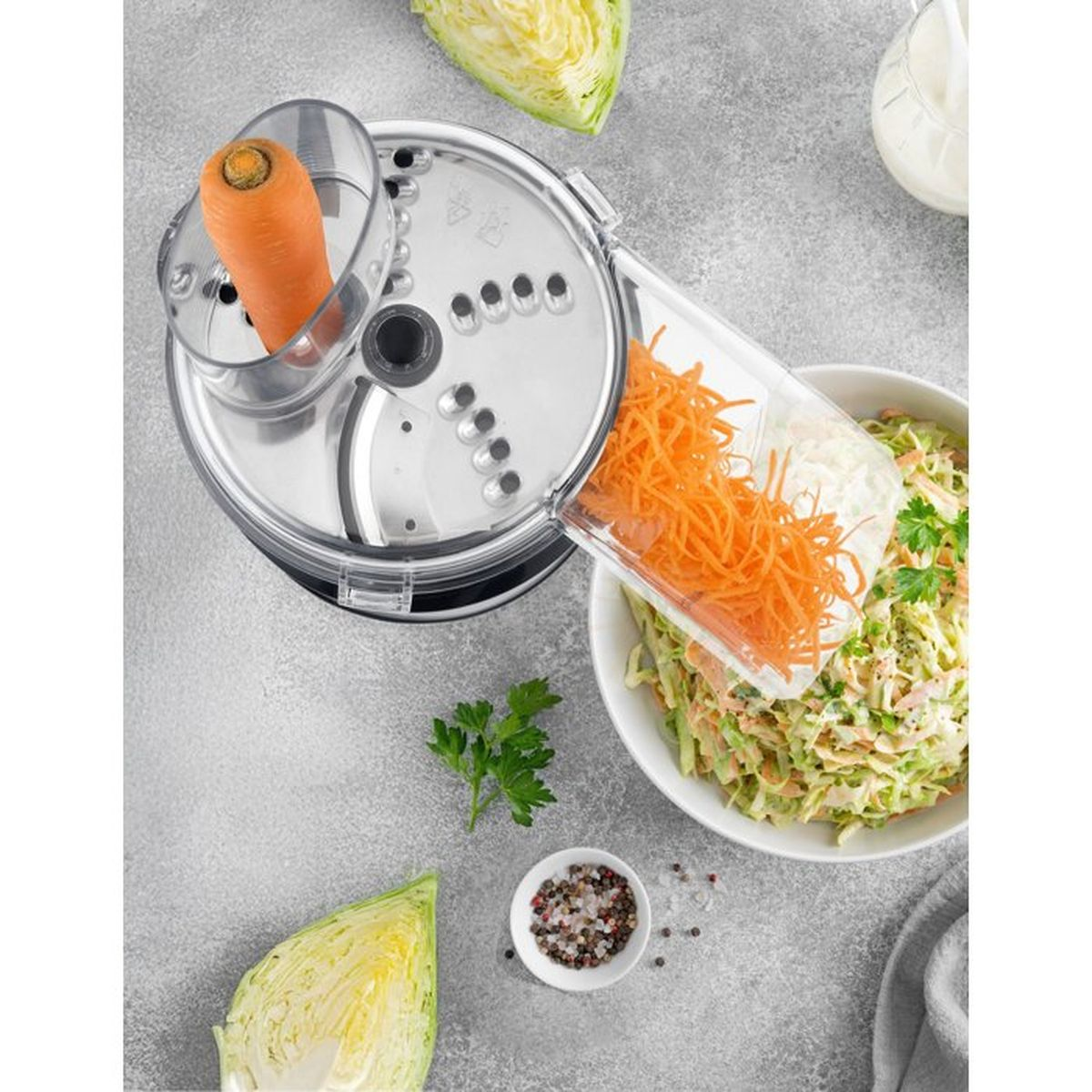 Un robot da cucina grattugia le carote in una ciotola di insalata, circondato da cavolo e spezie su un piano grigio.