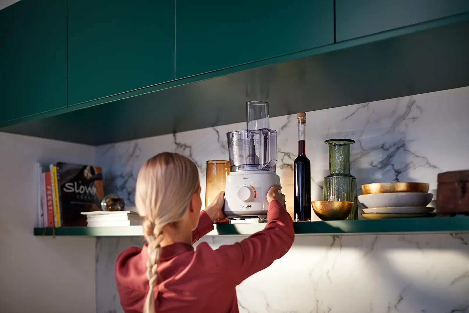 Eine blonde Frau im roten Hemd stellt eine weiße Philips Küchenmaschine in ein Regal.