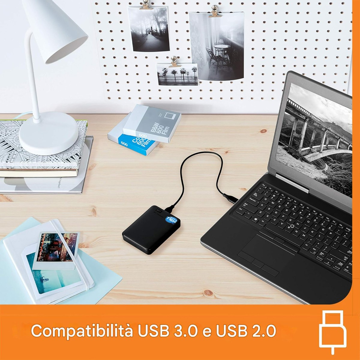 Un disco rigido esterno è collegato tramite cavo a un laptop su una scrivania di legno, con lampada e foto.