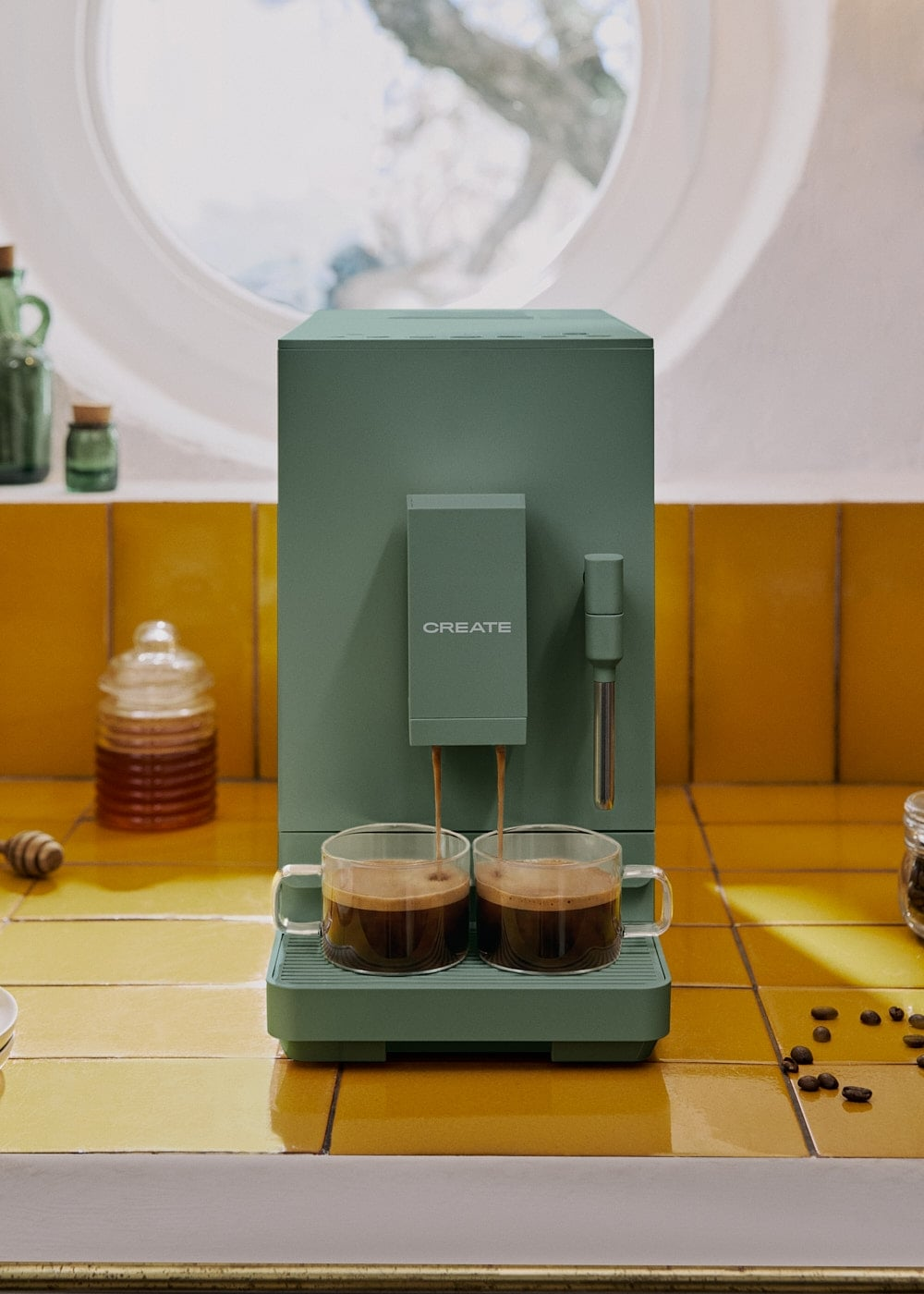 Una cafetera verde Create vierte café en dos tazas de vidrio sobre una encimera de azulejos amarillos.