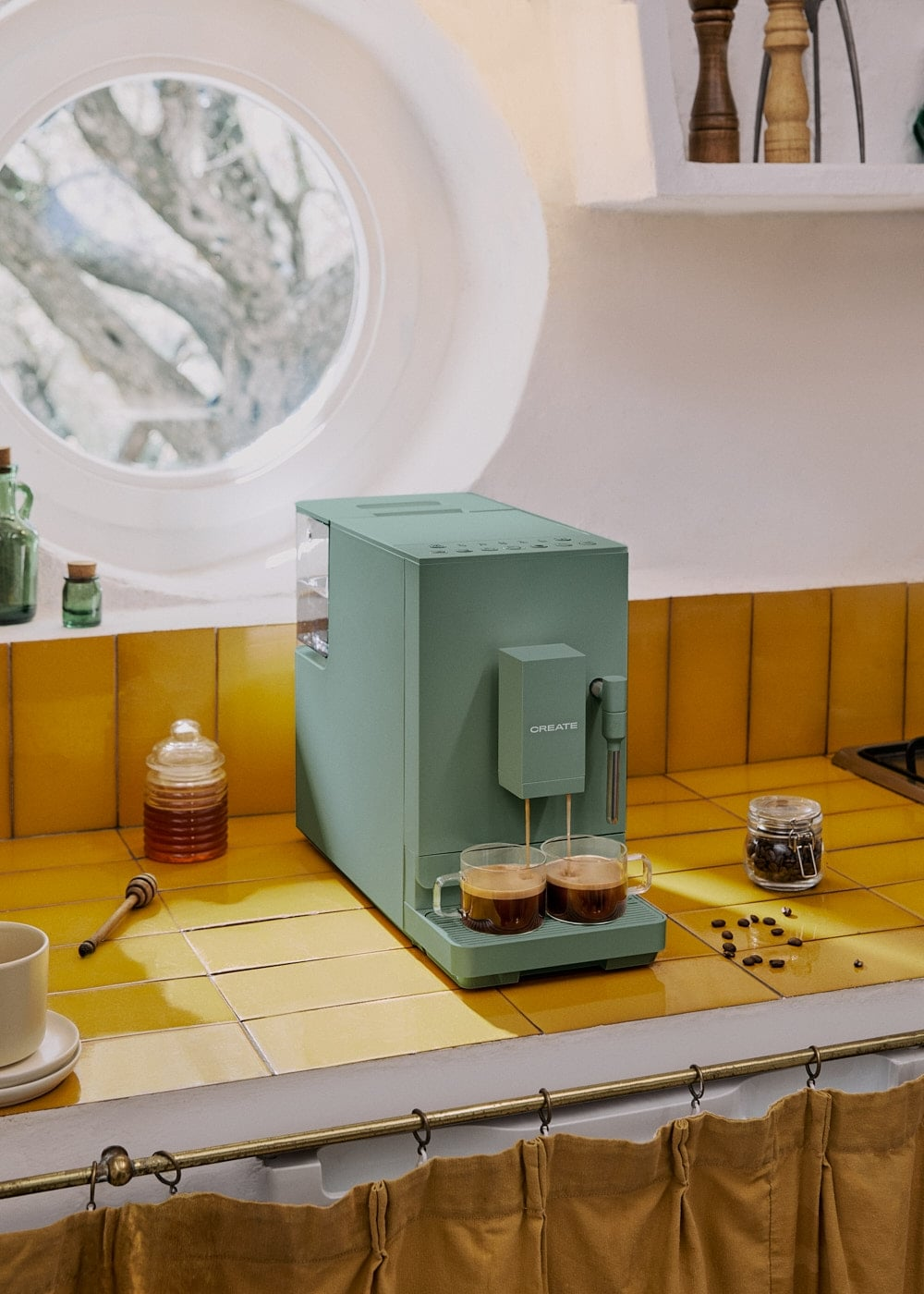 Una cafetera verde en una encimera de azulejos amarillos vierte café en dos tazas de vidrio, con una ventana redonda al fondo.