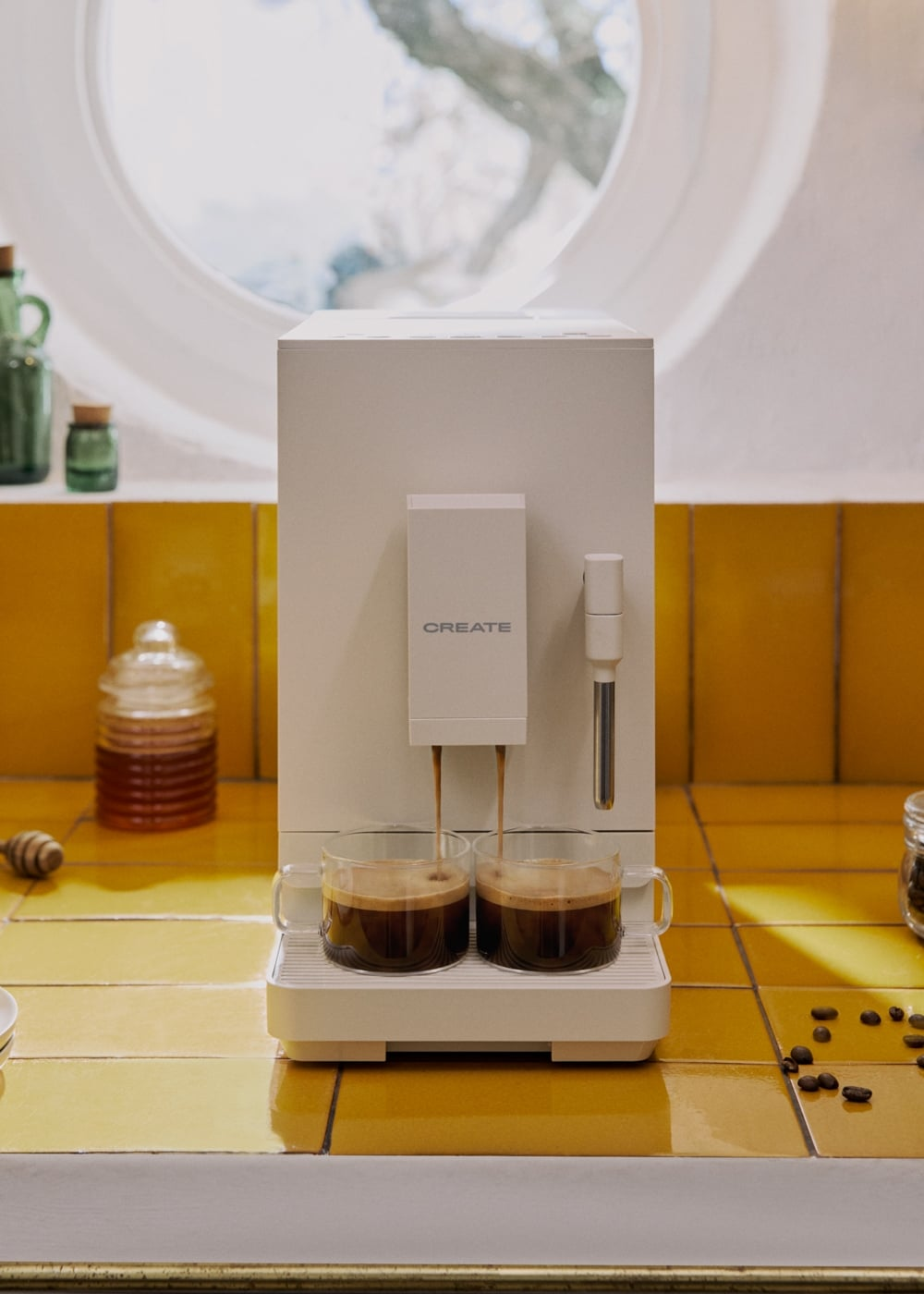 Una cafetera blanca vierte café en dos tazas de vidrio en un mostrador de azulejos amarillos.