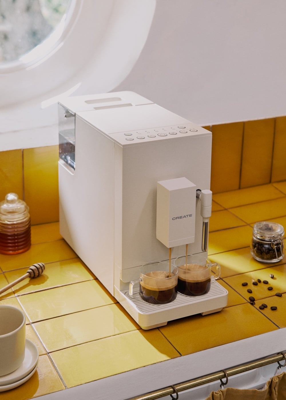 Una cafetera blanca vierte café en dos tazas de vidrio en un mostrador de azulejos amarillos.