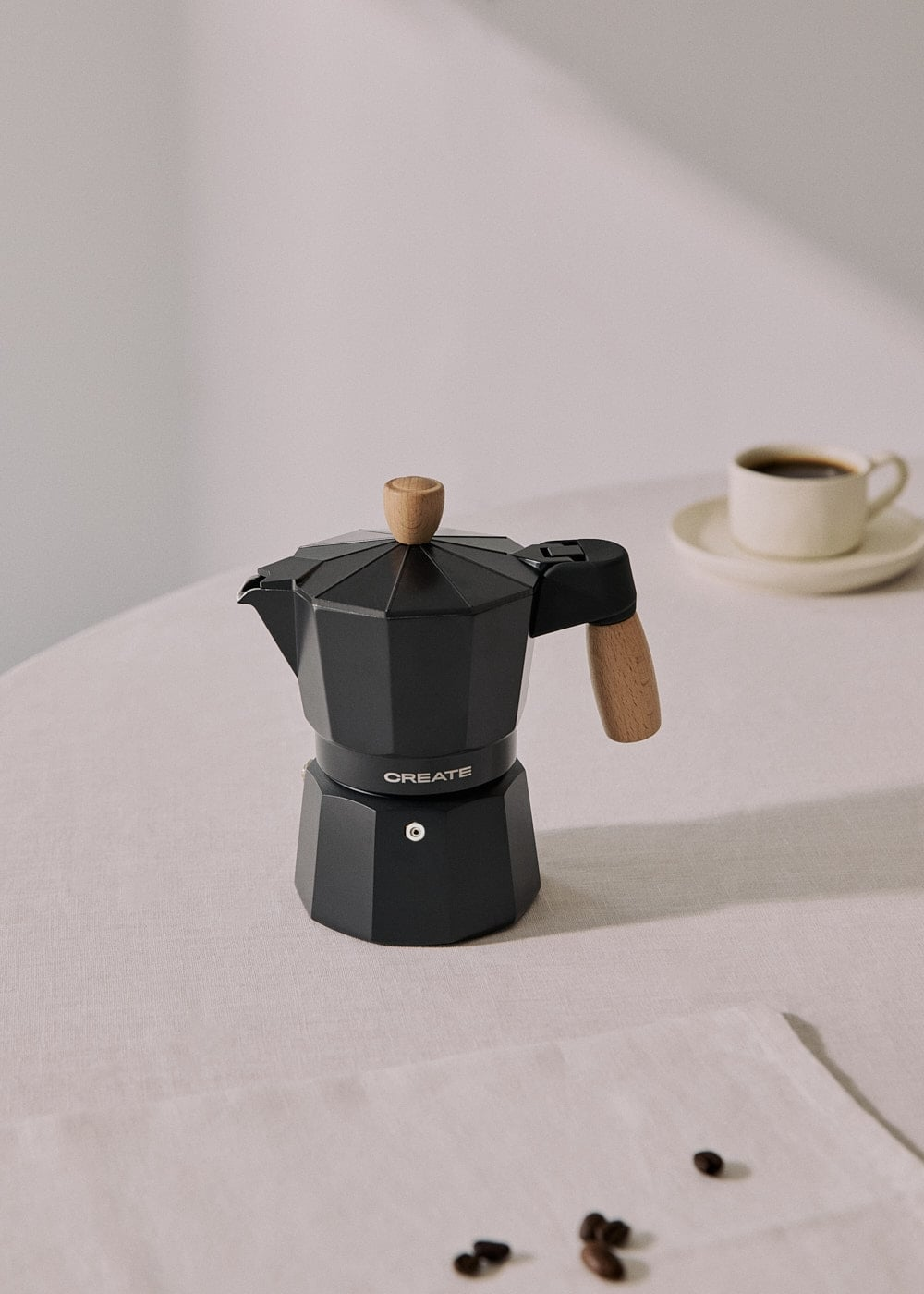 Una cafetera italiana negra con detalles de madera sobre una mesa junto a una taza y granos.