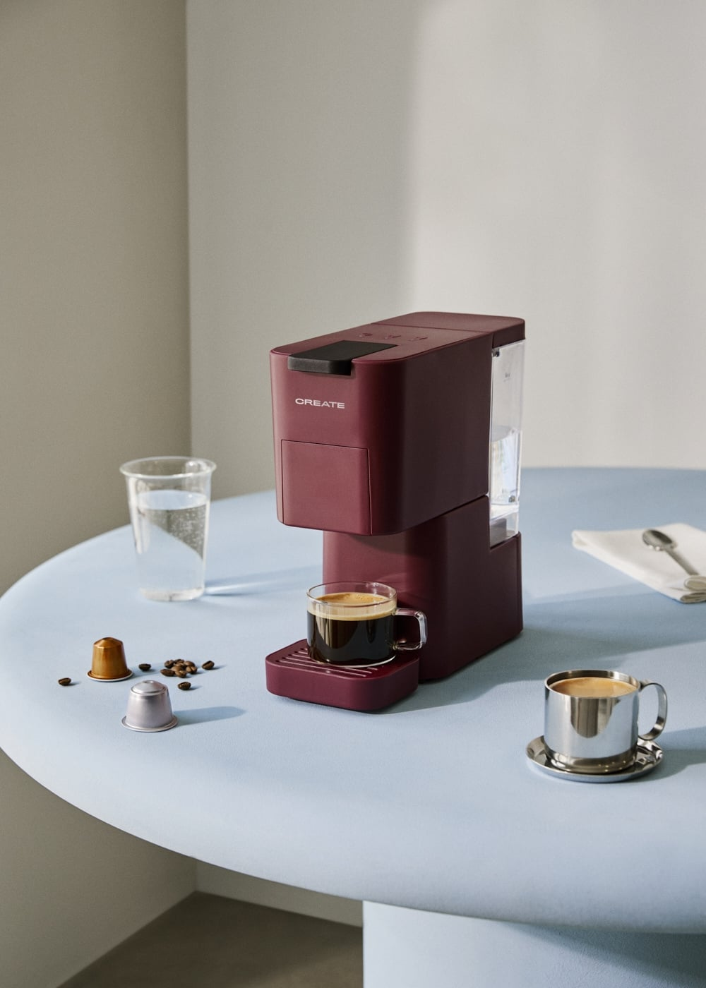 Una cafetera granate sobre una mesa redonda azul, con tazas de café y granos.