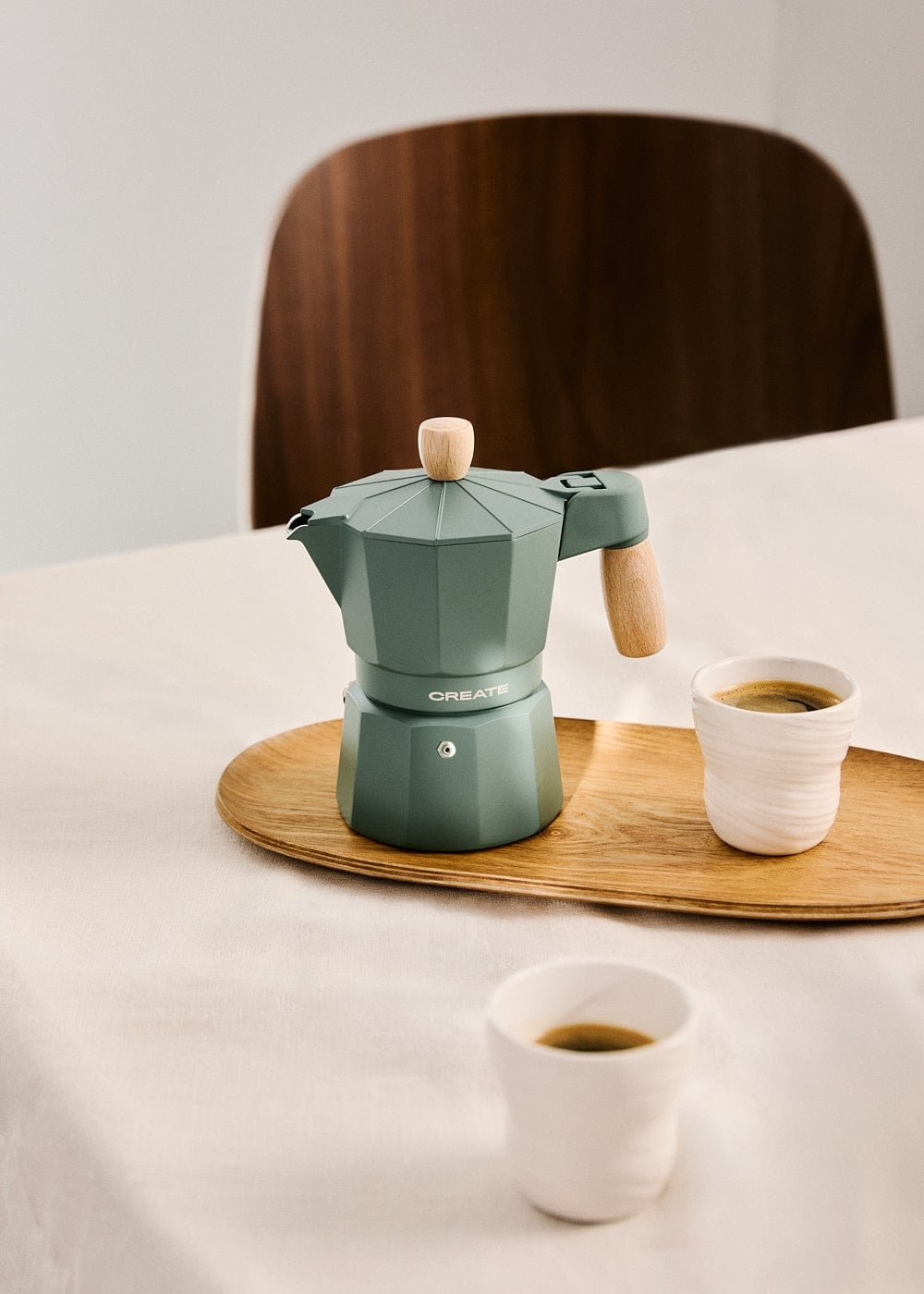 Una cafetera moka verde con asas de madera en una bandeja de madera con dos tazas de café.