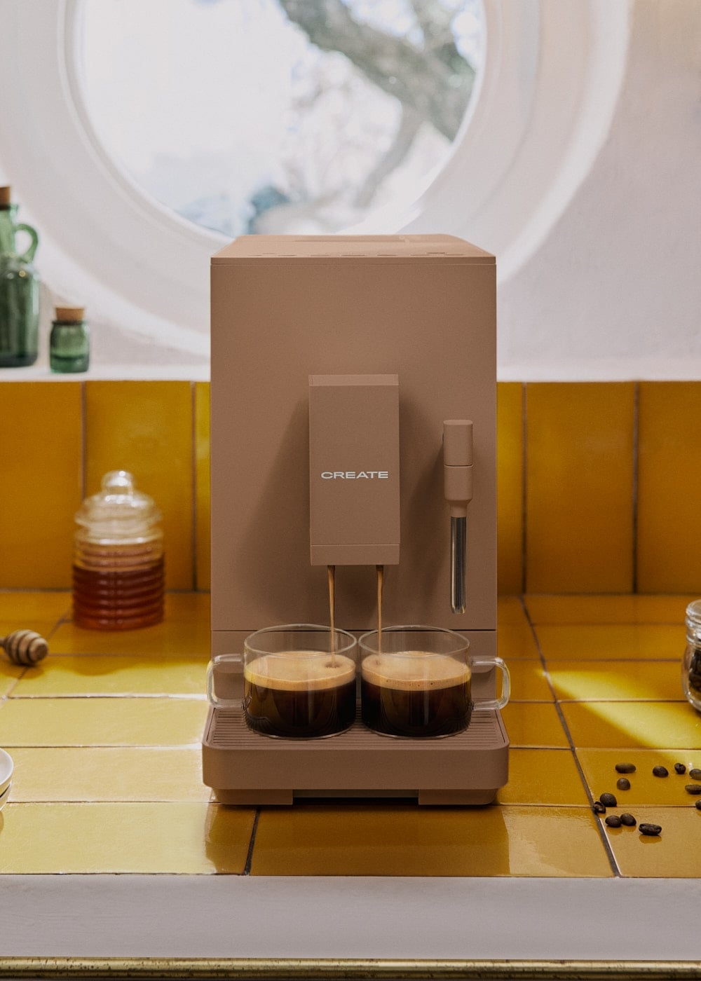 Una cafetera Create beis llena dos tazas de cristal en una encimera de azulejos amarillos.
