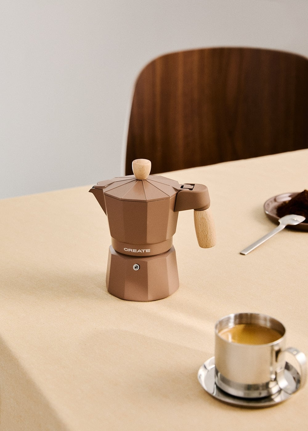 Una cafetera moka marrón con detalles de madera sobre un mantel beis cerca de una taza de metal.