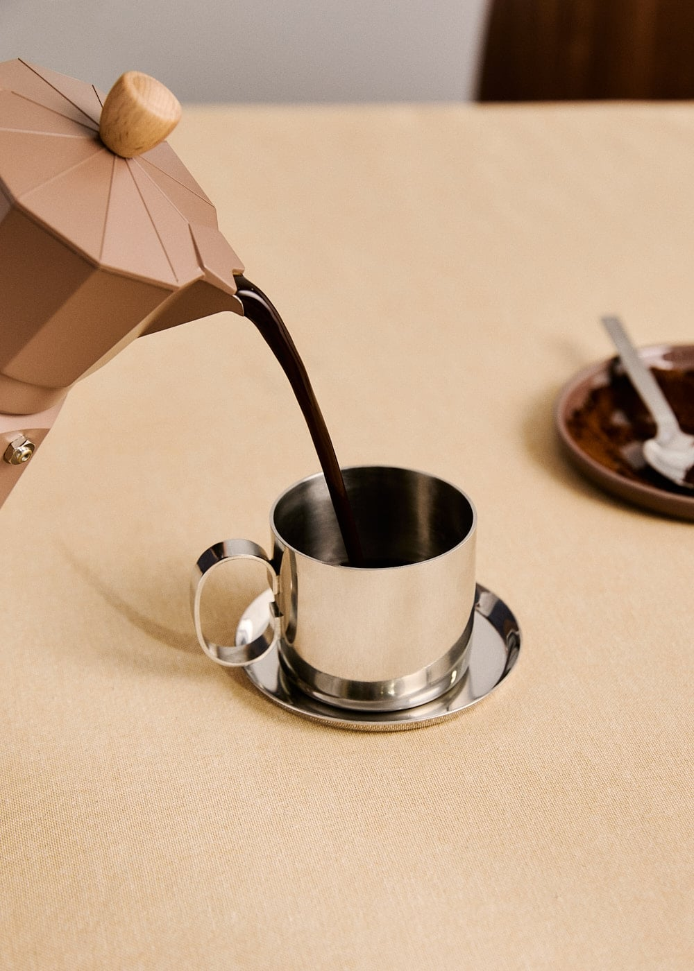 Una cafetera moka beige vierte café en una taza plateada con platillo en una mesa clara.