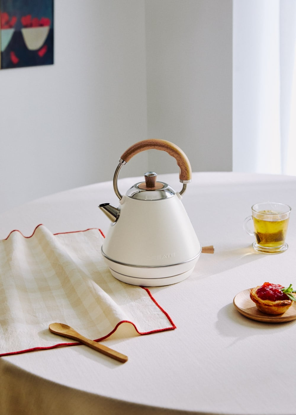Un hervidor blanco con asa de madera sobre una mesa junto a una taza de té y un pastel.