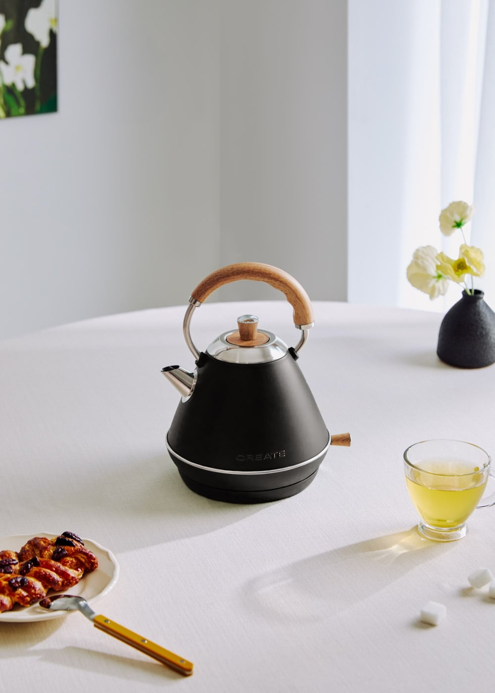 Un hervidor negro con asa de madera sobre una mesa puesta con pasteles y té.