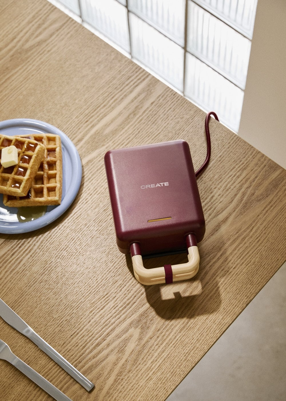 Una wafflera burdeos con asas de madera en una mesa de madera, junto a un plato de waffles.
