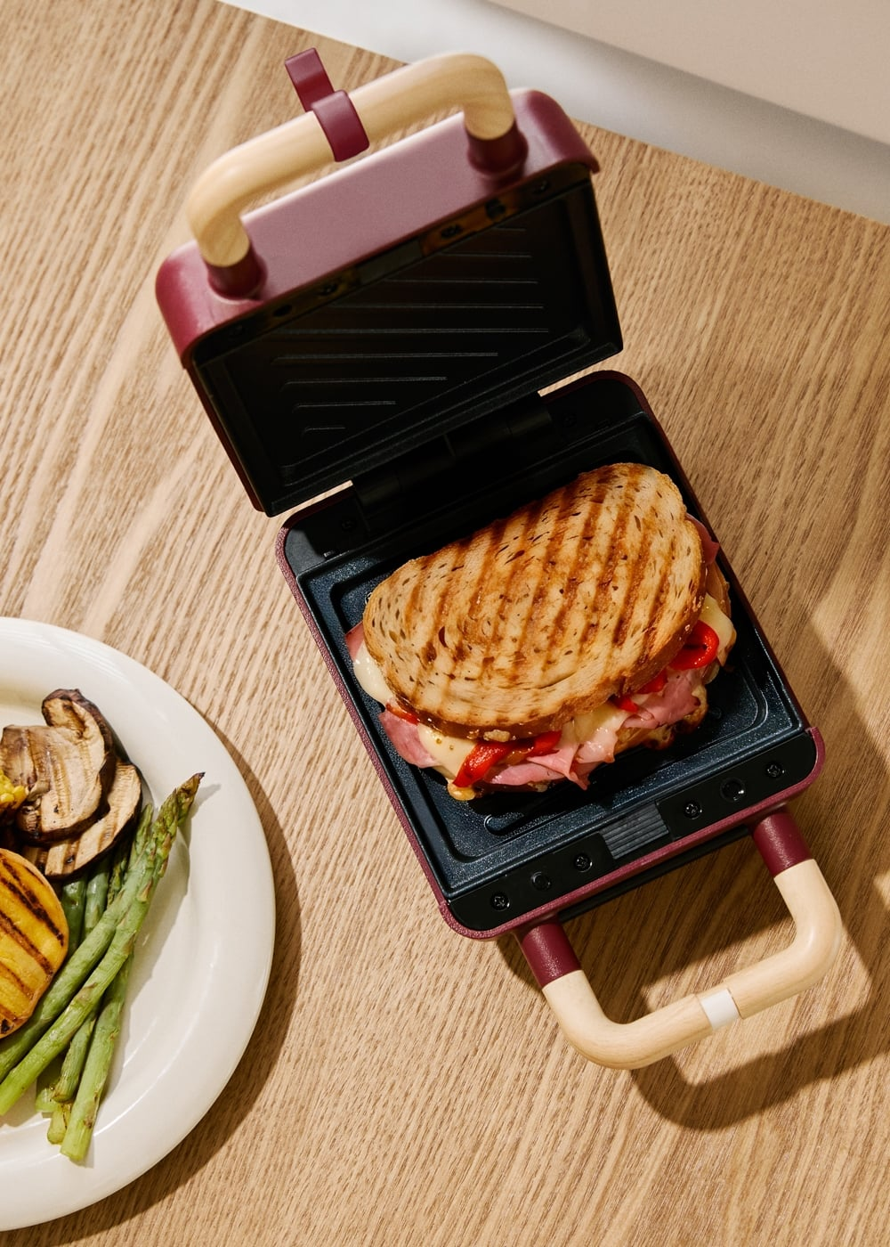 Un sándwich en una sandwichera roja abierta sobre una mesa de madera, con un plato de verduras.