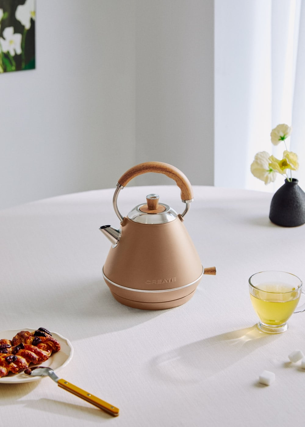 Un hervidor beige con asa de madera está sobre una mesa con repostería, té y flores.
