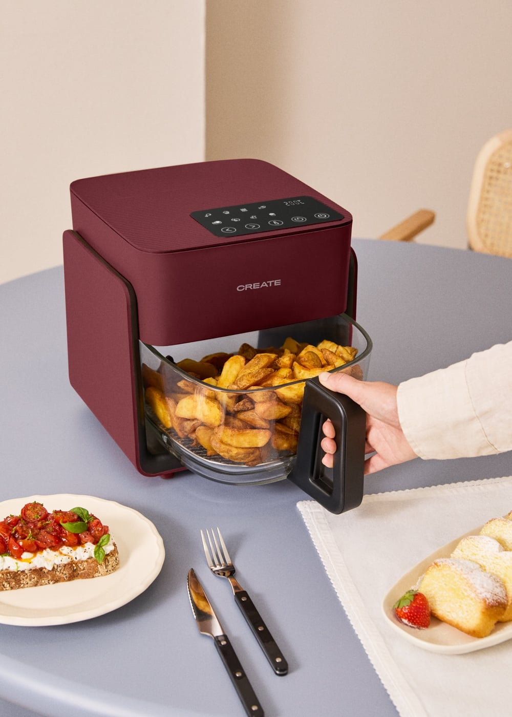 Una mano tira de la cesta de una freidora de aire roja llena de patatas fritas. Hay tostadas y pasteles en la mesa.