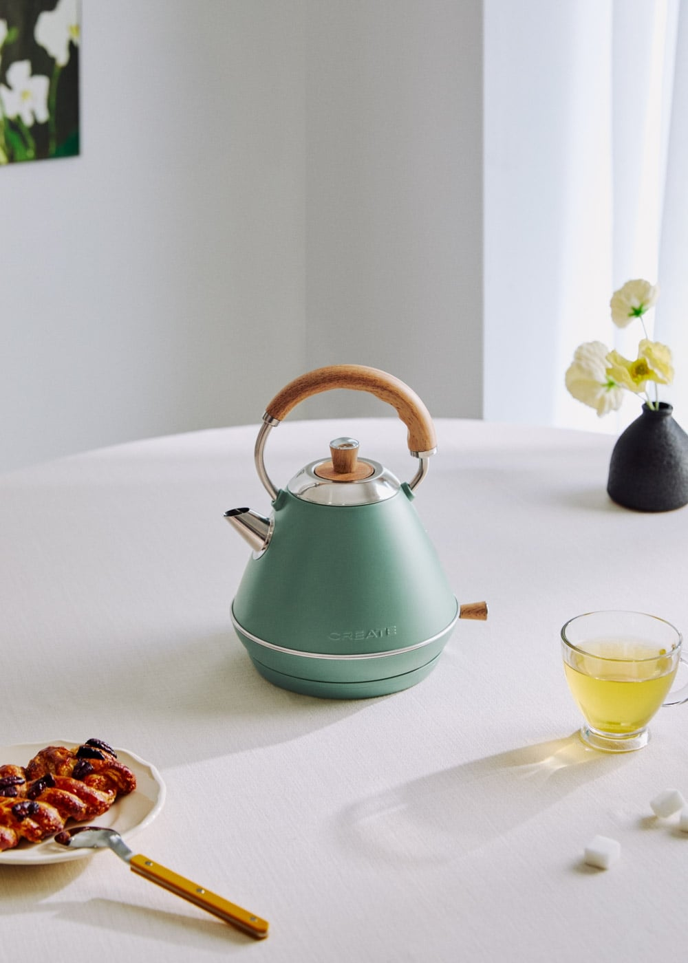 Un hervidor eléctrico verde pastel con asa de madera sobre un mantel blanco, acompañado de té y bollería.
