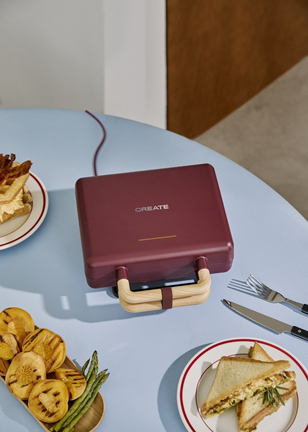 Una sandwichera burdeos con mangos de madera en una mesa azul, rodeada de platos de comida.