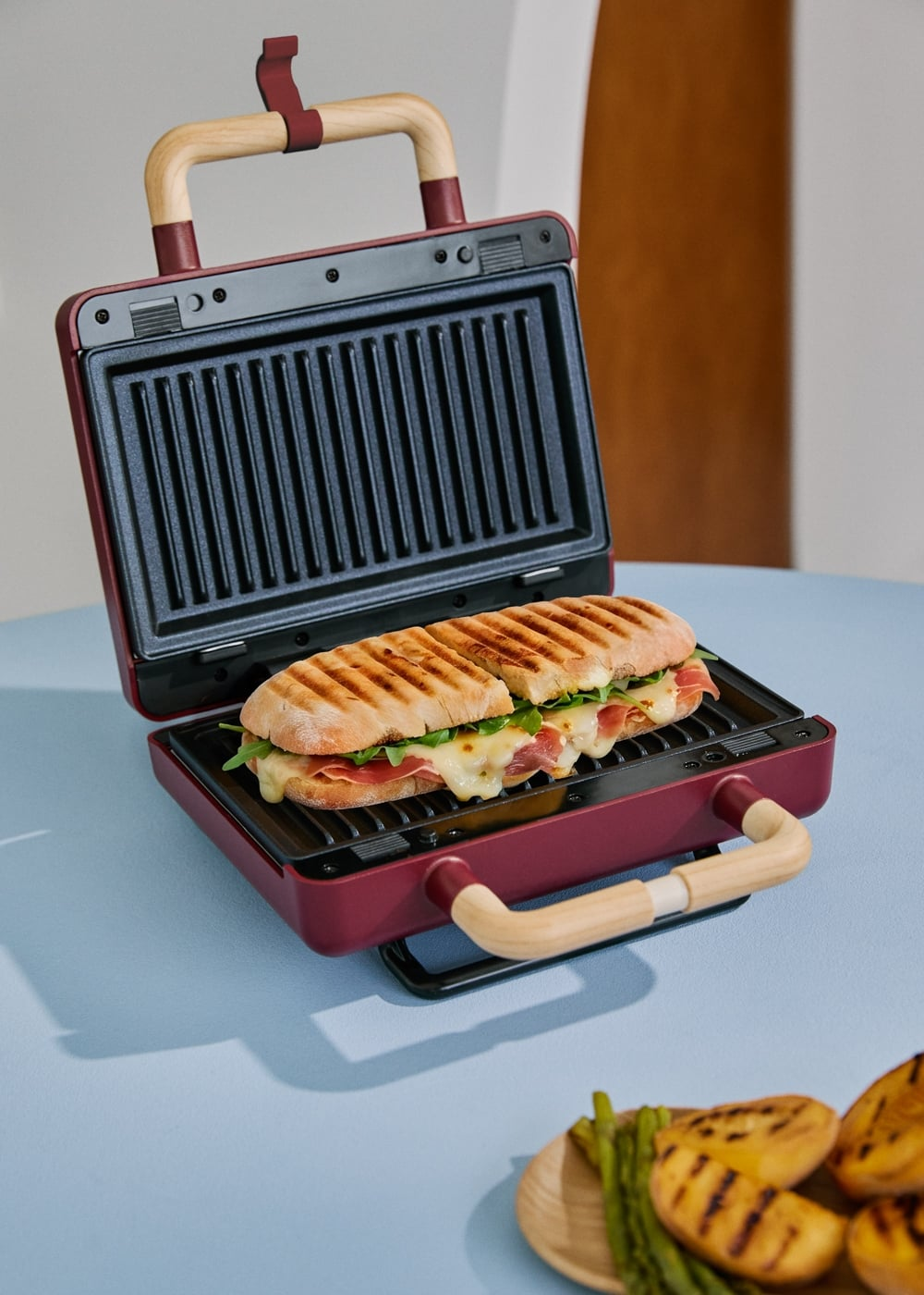 Una prensa para panini está abierta en una mesa azul, con un panini a la parrilla con queso derretido, jamón y verduras.