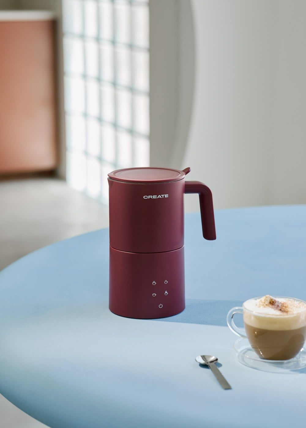 Un espumador de leche rojo sobre una mesa azul junto a una taza de capuchino y una cuchara.