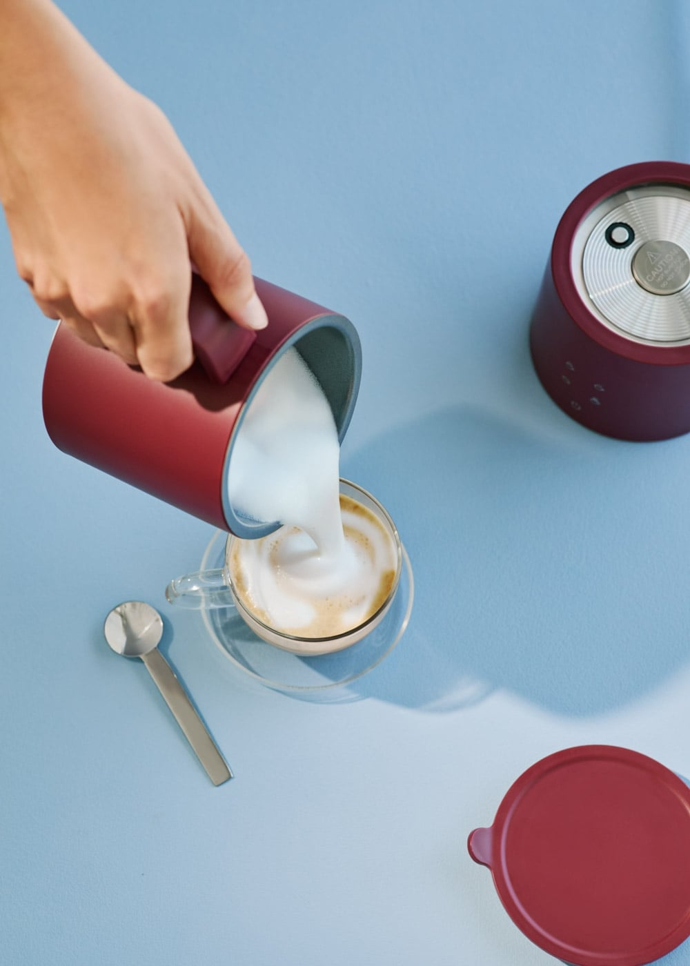 Se vierte espuma de leche de una jarra roja en una taza de café sobre una mesa azul.