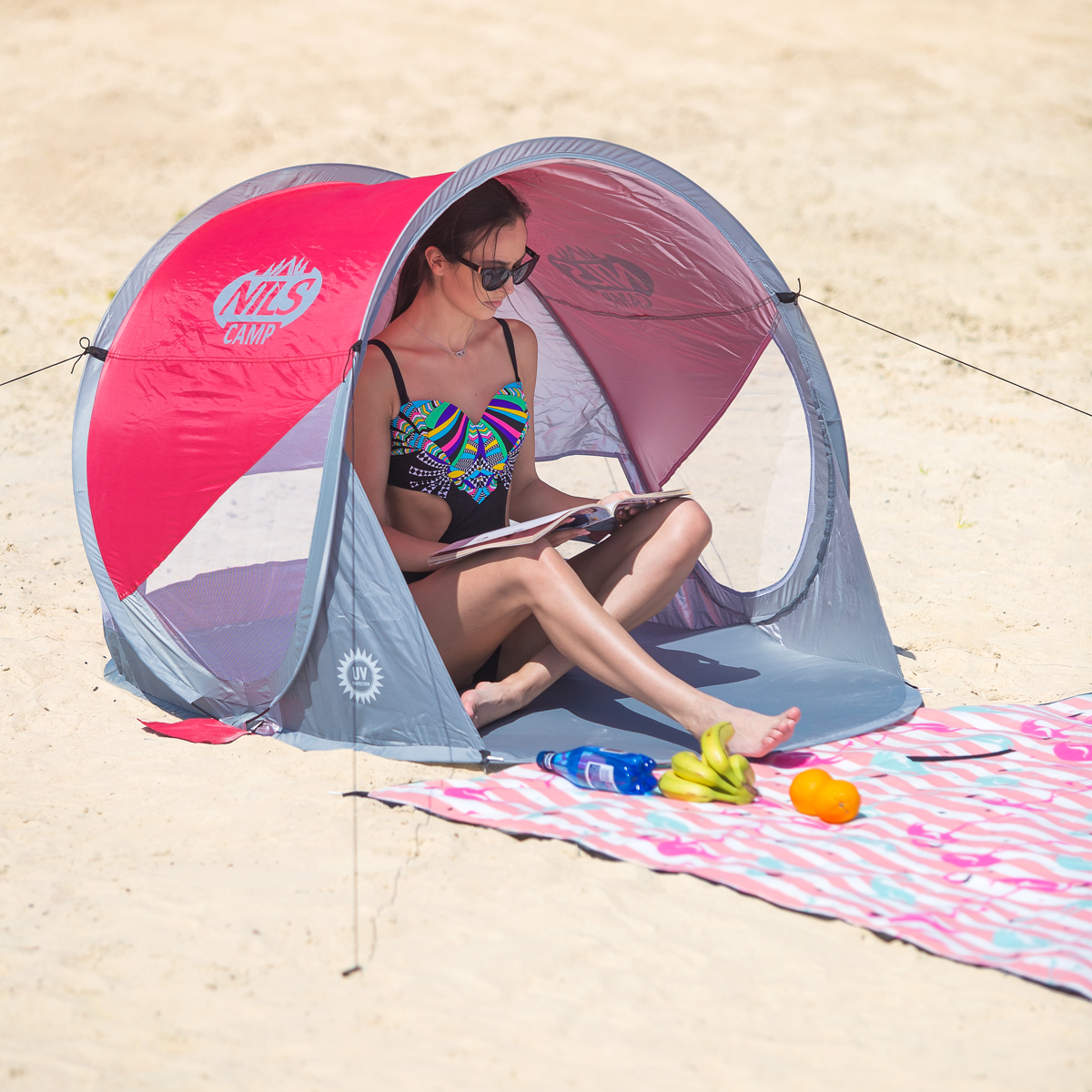 Kobieta w kolorowym stroju kąpielowym czyta książkę w czerwono-szarym namiocie plażowym.