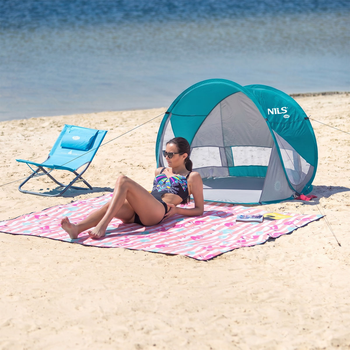 Kobieta w stroju kąpielowym relaksuje się na kocu plażowym obok niebieskiego namiotu i krzesła.