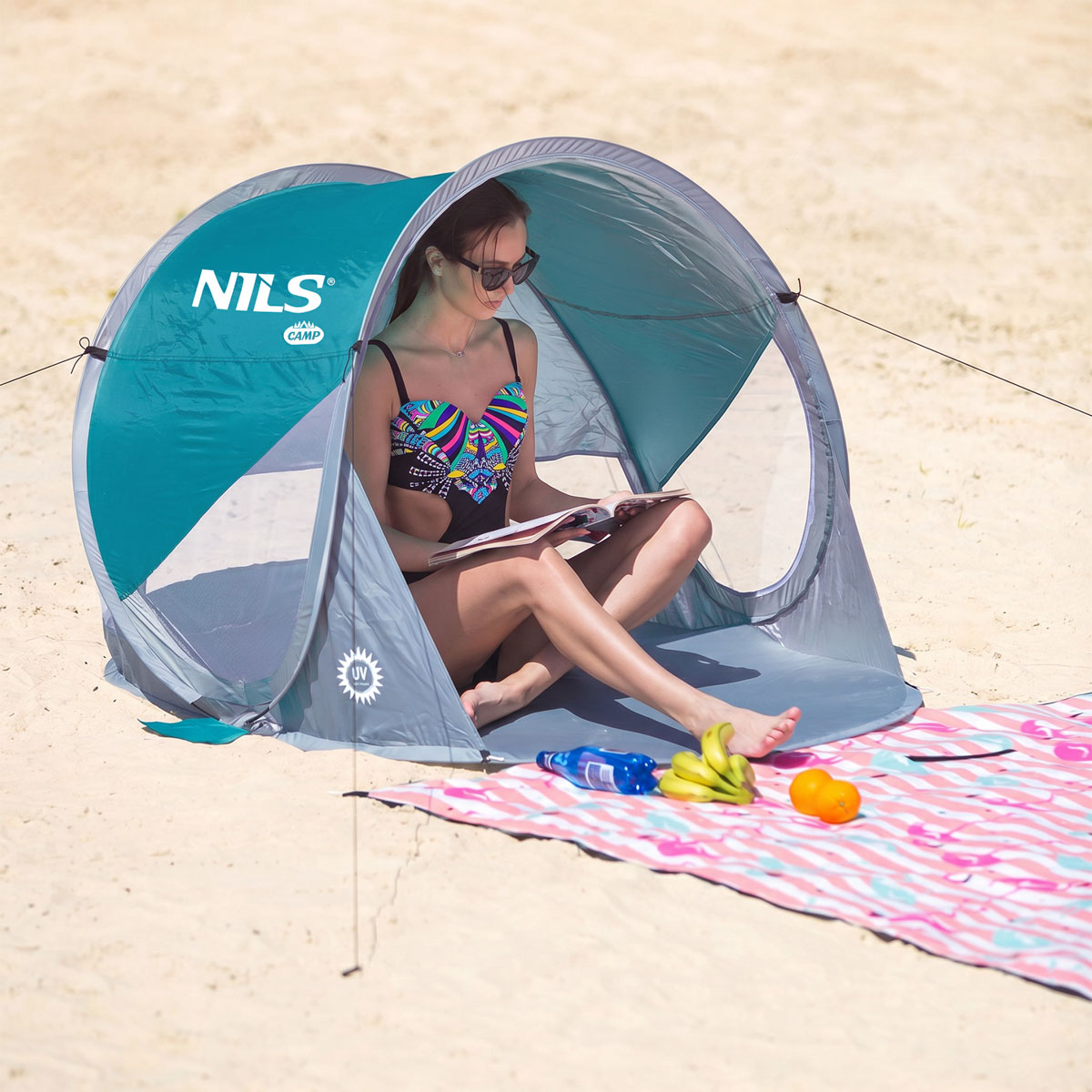 Kobieta w stroju kąpielowym czyta książkę w niebieskim namiocie plażowym na piasku.
