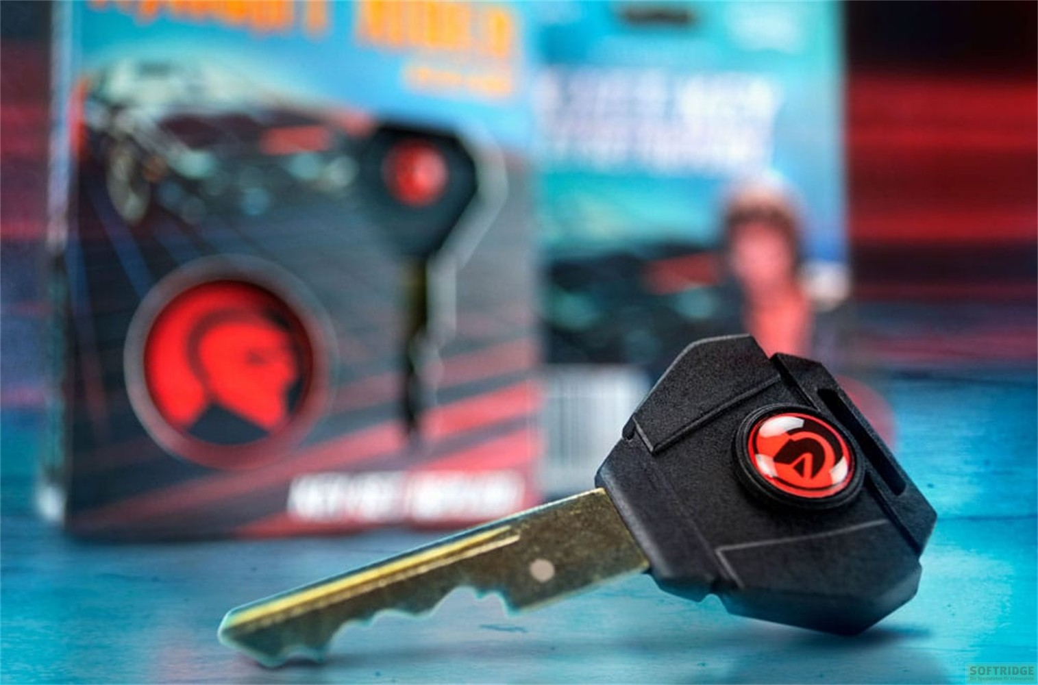 Una llave de coche negra con un logo rojo yace sobre una superficie azul frente a un embalaje.