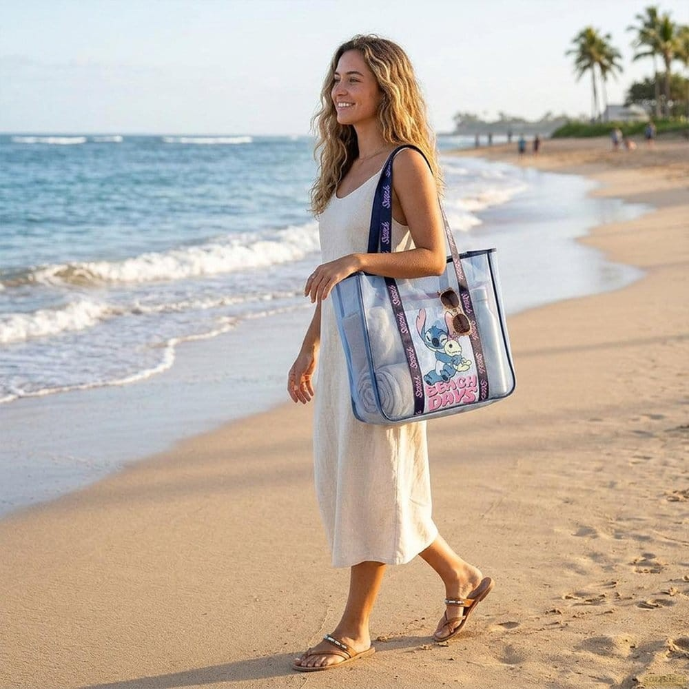 Una mujer rubia con vestido claro camina por la playa llevando un bolso transparente con un dibujo animado.