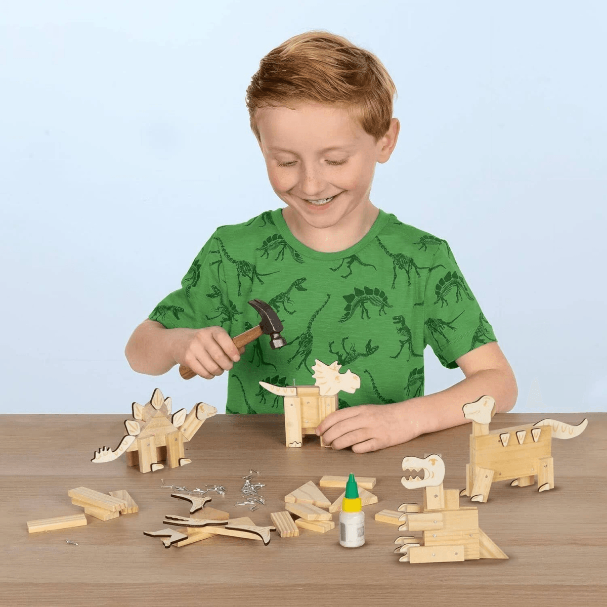 Un niño construye dinosaurios de madera en una mesa con martillo y pegamento.