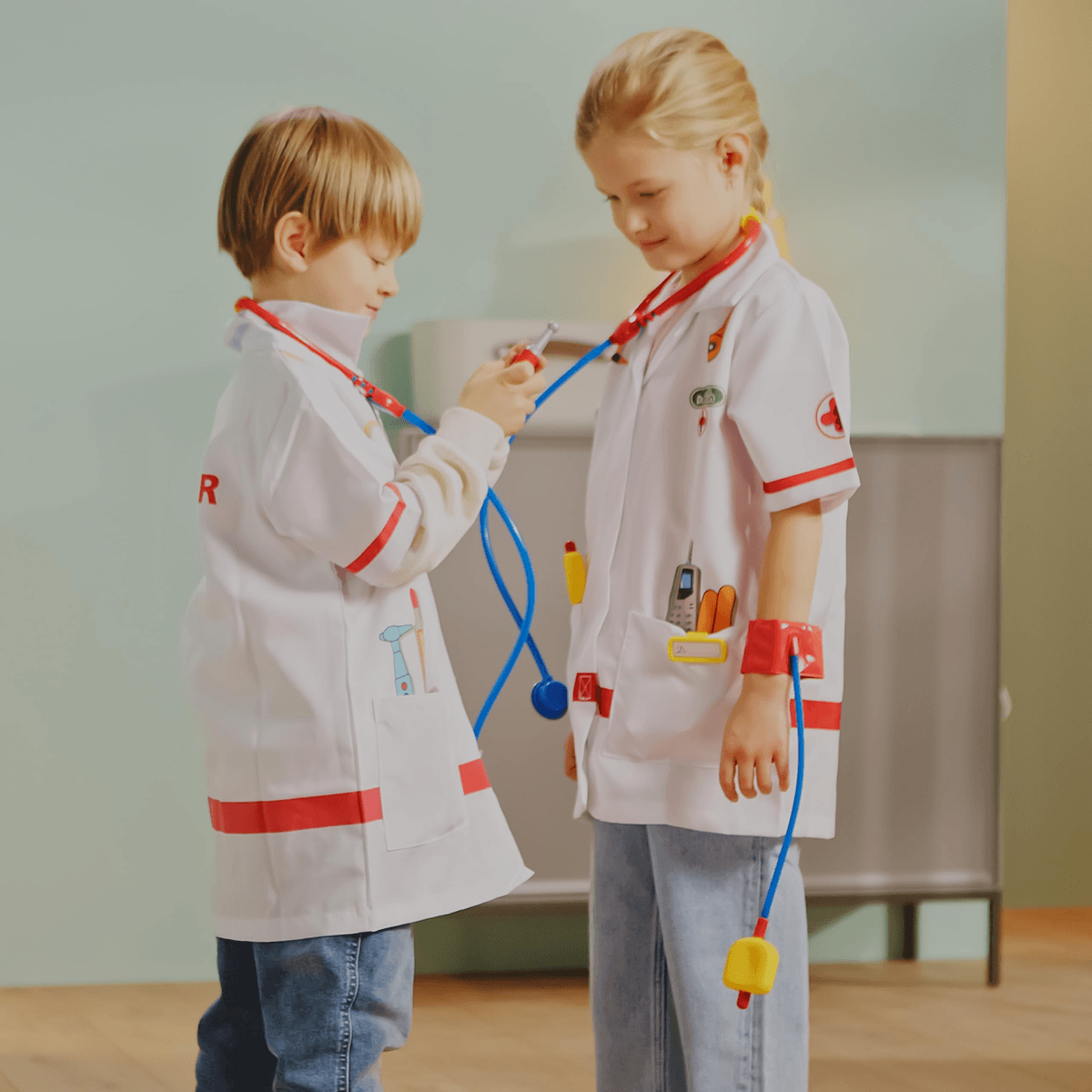 Dos niños con batas de médico juegan con estetoscopios en interior.