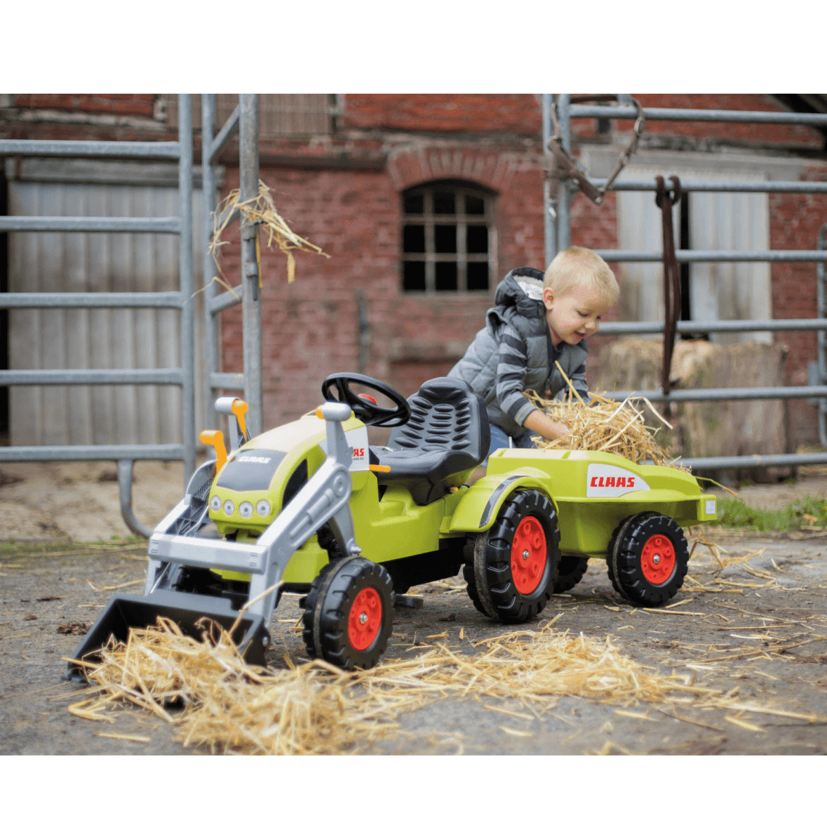 Un niño pequeño juega con un tractor de juguete verde con remolque, cargando heno en una granja.