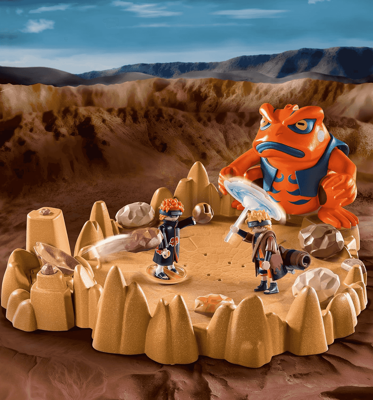 Figuras de acción de Naruto luchando en una arena rocosa con un gran sapo naranja al fondo.